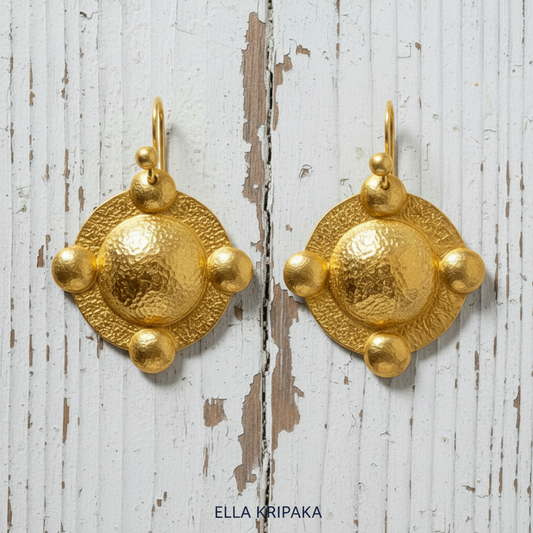 Pair of hammered 24k gold convex textured earrings with convex rounds on a old white wooden background, and with 'Ella Kripaka' branding.