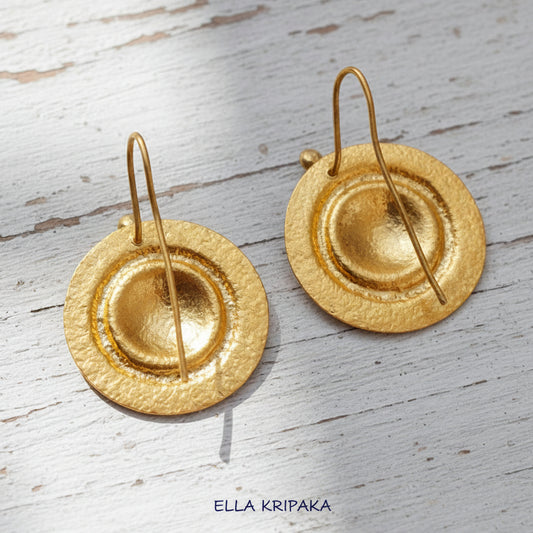 Pair of hammered 24k gold convex disks earrings on the reverse side and with hook clasps on a light wooden surface with 'Ella Kripaka' branding