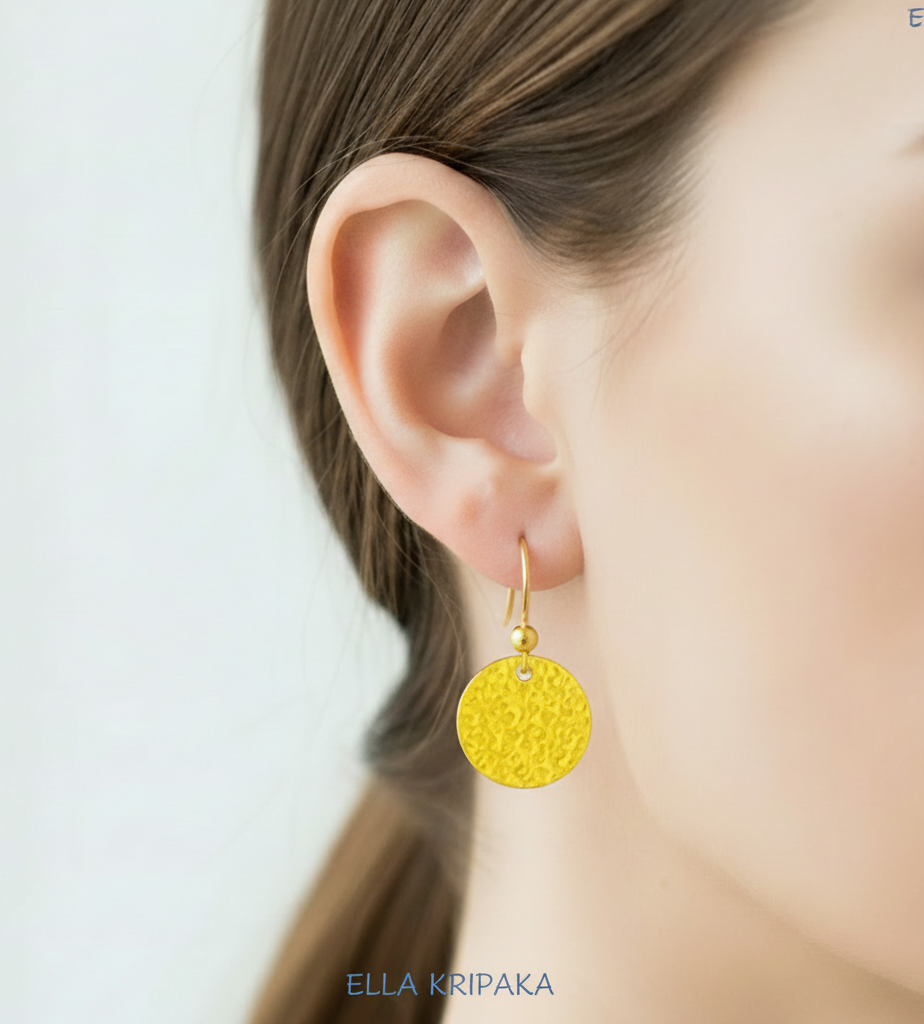 Hammered 24k gold, pure 9999 gold disk earring with shiny forged gold textures worn on a woman and with light grey background, artist-jeweler 'Ella Kripaka' branding