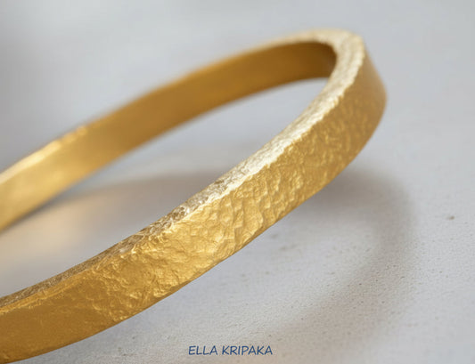 Thick hammered 24k gold shiny bracelet with forged textures on a light grey concrete surface, with artist 'ELLA KRIPAKA' branding