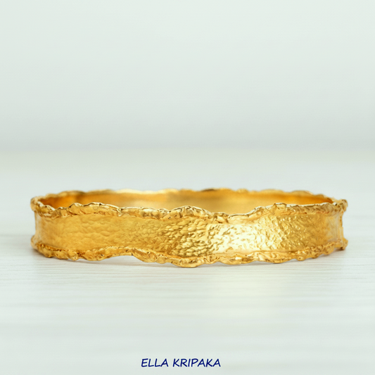 Organic Hammered 24k Gold Bangle Bracelet with uneven thick edges and shiny gold textures on a white wooden surface, branding artist-jeweler 'Ella Kripaka'