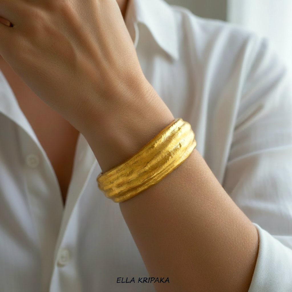 Organic 24k gold hammered textured bangle bracelet with convex uneven reliefs is worn by a model on hand and with blurred white shirt background, artist 'Ella Kripaka' branding