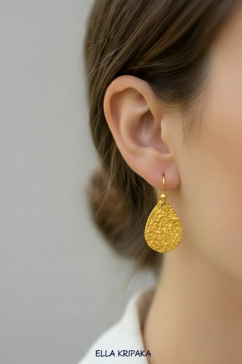 Hammered 24k gold textured drop earring with hook clasp is worn by a woman on grey background, with artist 'Ella Kripaka' branding