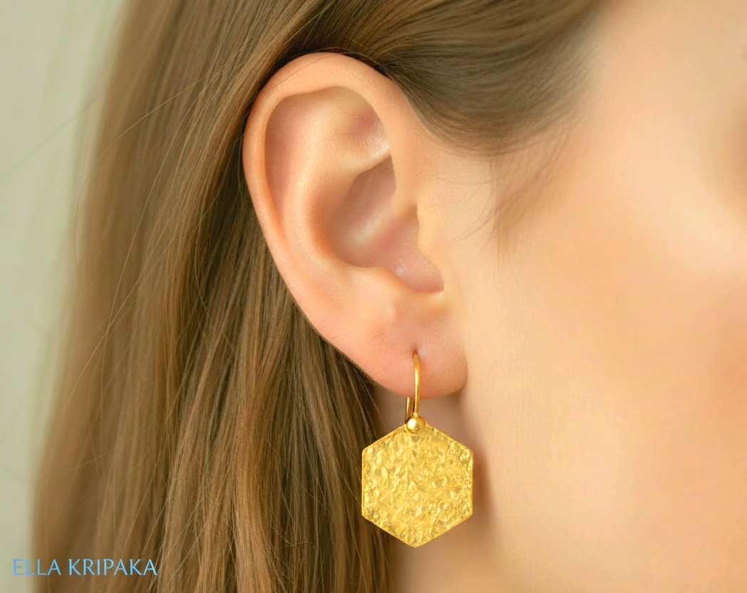 Hammered 24k gold, pure 9999 gold hexagon earring with shiny forged gold textures worn on a woman and with light grey background, artist-jeweler 'Ella Kripaka' branding