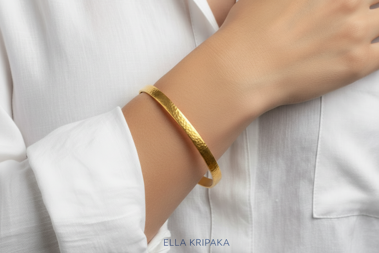 Hammered 22k gold bangle bracelet Ancient Rome style on model's wrist and on white blurred shirt background, with 'Ella Kripaka' branding