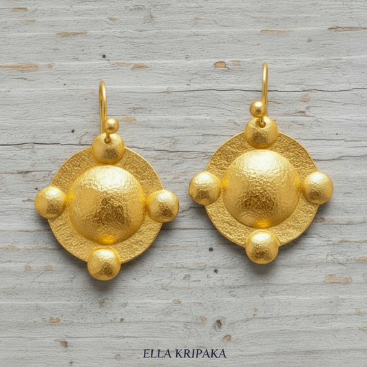 Pair of hammered 24k gold convex textured earrings with convex rounds on a rustic grey wooden background, and with 'Ella Kripaka' branding.
