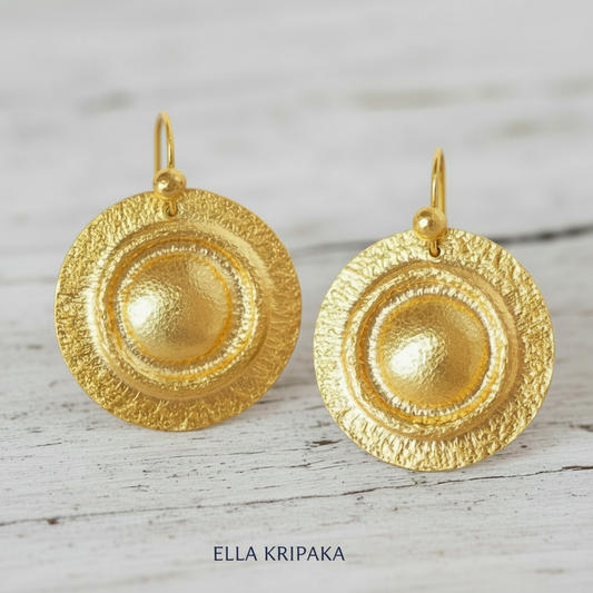 Pair of hammered 24k gold convex disks earrings with hook clasps on a light wooden surface with 'Ella Kripaka' branding