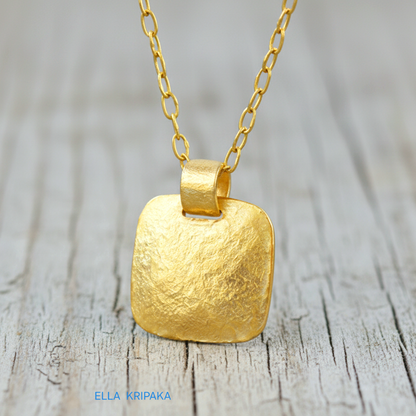 Hammered 24k gold textured convex square pendant with chain on light grey rustic wooden surface with 'Ella Kripaka' branding