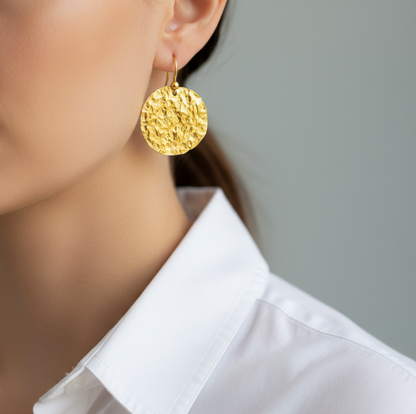 Organic 24k 9999 gold hammered disk uneven earrings with shiny gold textures are worn by a woman and on grey background, artist-jeweler's name 'Ella Kripaka'