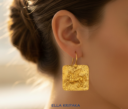 Hammered 24k 9999 gold square earring with convex Pegasus worn by a person with a blurred background, artist's name 'Ella Kripaka'