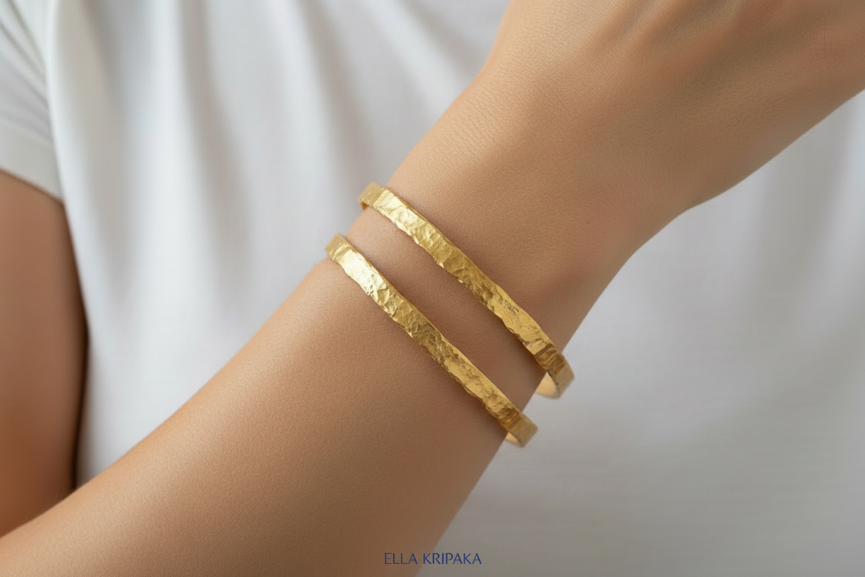 Organic hammered 24k 9999 Gold two bangles uneven bracelets worn by a person on wrist and with white t-shirt background, artist's name 'Ella Kripaka' branding