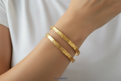 Organic hammered 24k 9999 Gold two bangles uneven bracelets worn by a person on wrist and with white t-shirt background, artist's name 'Ella Kripaka' branding