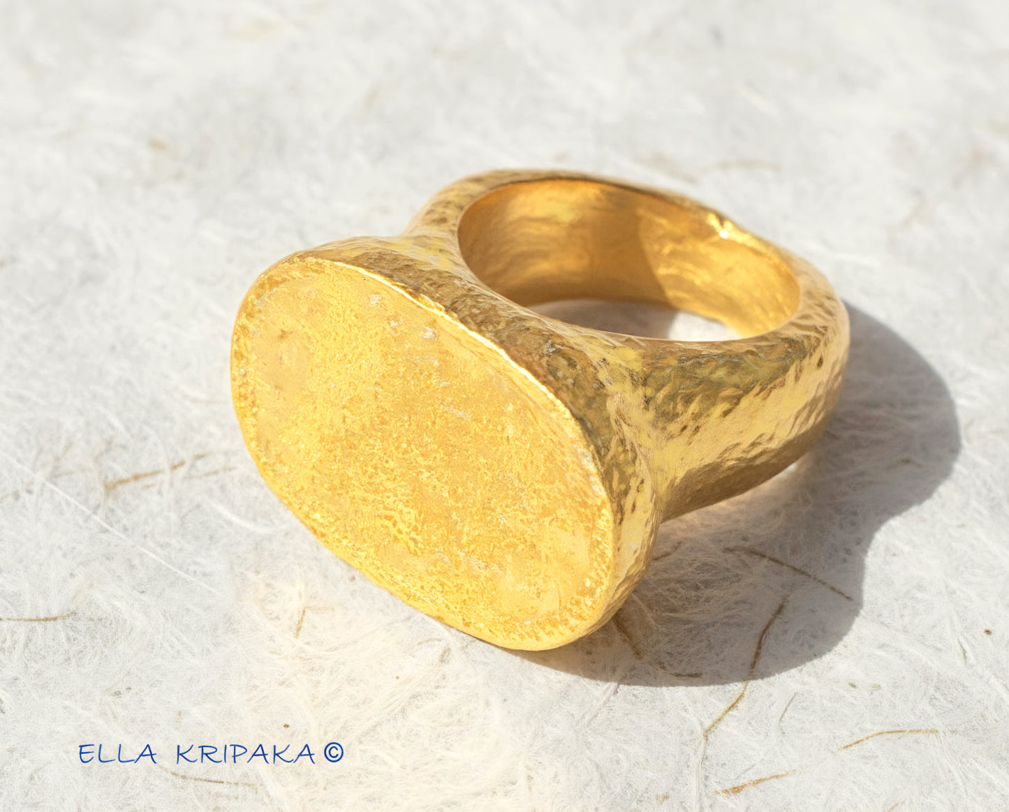 Hammered 24k 9999 Gold ring thick signet Ancient Egypt style and Ancient Rome style and with forged textured gold on a white paper surface, artist-jeweler's name 'Ella Kripaka' branding