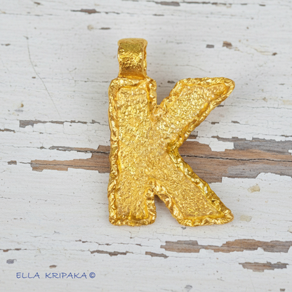 24k 9999 Gold hammeted letter Initial 'K' pendant on a textured wooden background, artist's name 'Ella Kripaka'