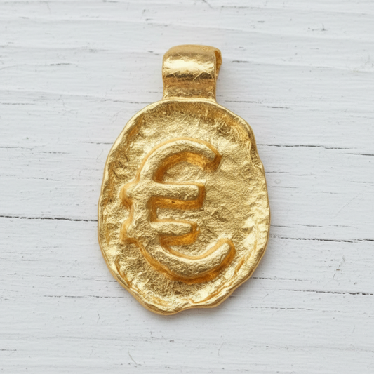 Organic 24k gold hammered uneven pendant with convex 'Euro' sign on a old wooden surface, branded artist 'Ella Kripaka'