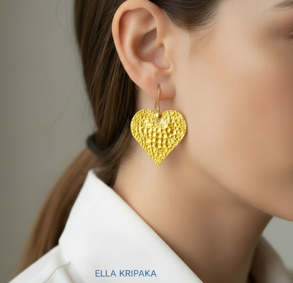 Organic 24k 9999 gold hammered heart earrings with shiny gold textures are worn by a model on neutral background, artist-jeweler's name 'Ella Kripaka'