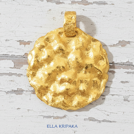 Organic 24k 9999 gold hammered uneven disk pendant on a white wooden surface, branded 'Ella Kripaka'