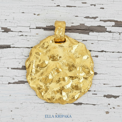 Organic 24k 9999 gold hammered uneven disk pendant on a wooden surface, branded 'Ella Kripaka'