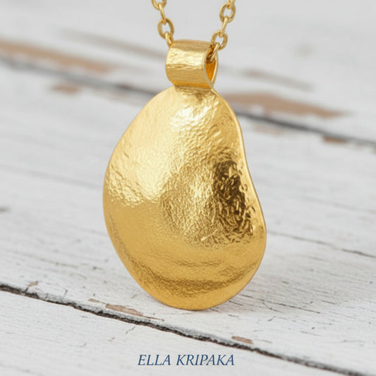 Organic 24k gold convex uneven oval pendant with hammered textures anx with chain is on old white wooden surface, branding artist 'Ella Kripaka'