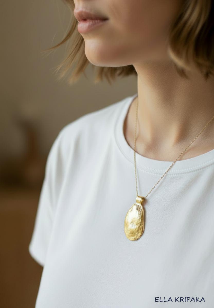 Organic 24k gold convex uneven oval pendant with hammered textures and on chain is worn by a woman with white t-shirt and on neutral background, branding artist 'Ella Kripaka'