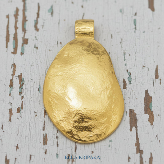 Organic 24k gold convex uneven oval pendant with hammered textures is on old white wooden surface, branding artist 'Ella Kripaka'