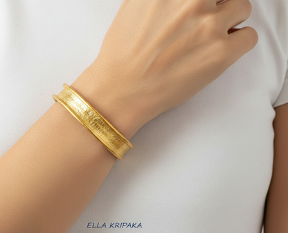 Hammered 24k 9999 gold bracelet cuff with thuck borders is worn on a woman's wrist and on white blurred background, branding artist-jeweler name 'Ella Kripaka'