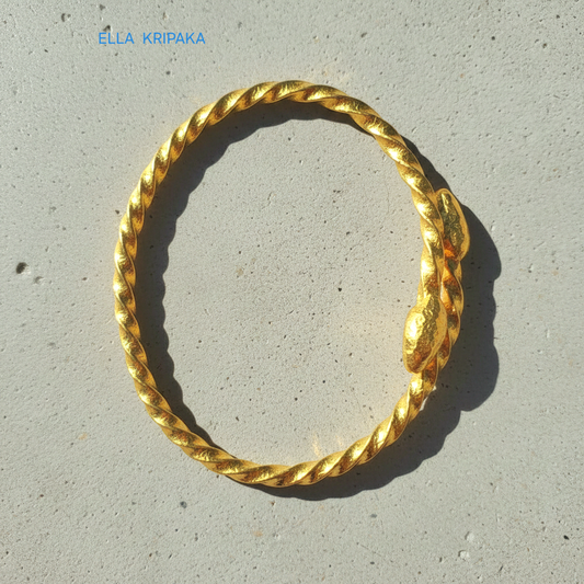 Twisted 24k gold hammered bracelet with thick oval ends and with textured design on grey concrete surface, branded artist-jeweler 'Ella Kripaka'