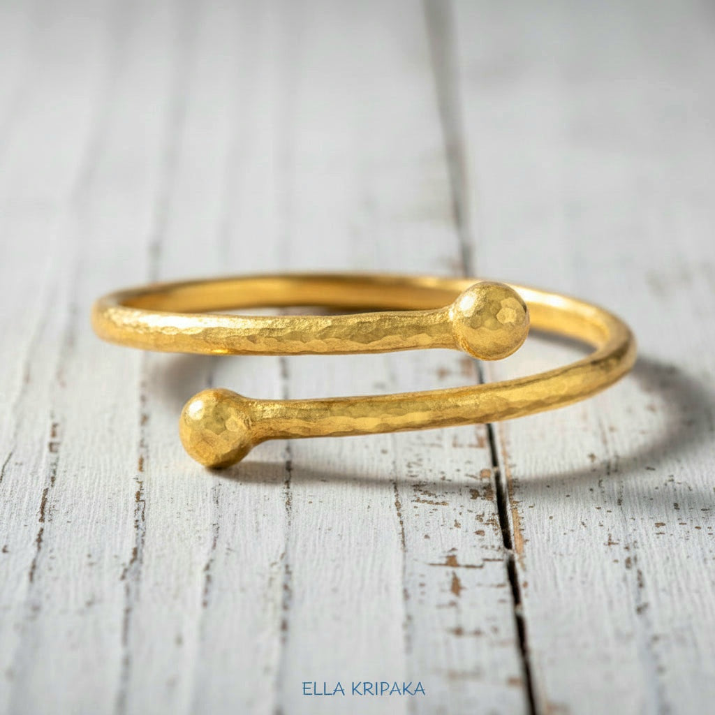 Twisted 24k gold hammered bracelet with thick convex rounds ends and with textured design on white rustic wooden surface, branded artist-jeweler 'Ella Kripaka'