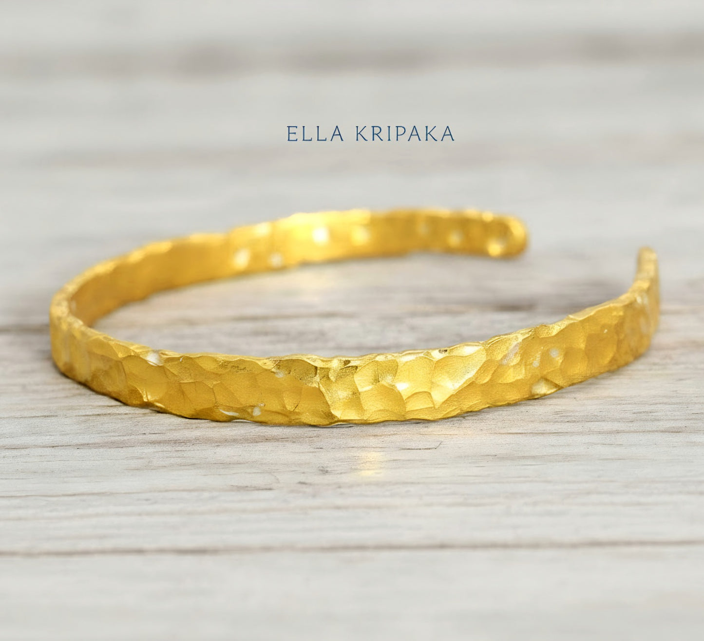 Organic hammered 24k gold bracelet cuff with uneven edges and textured design is on light wooden surface, branding artist's name 'Ella Kripaka'