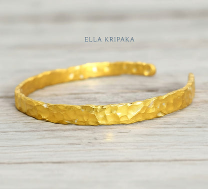 Organic hammered 24k gold bracelet cuff with uneven edges and textured design is on light wooden surface, branding artist's name 'Ella Kripaka'