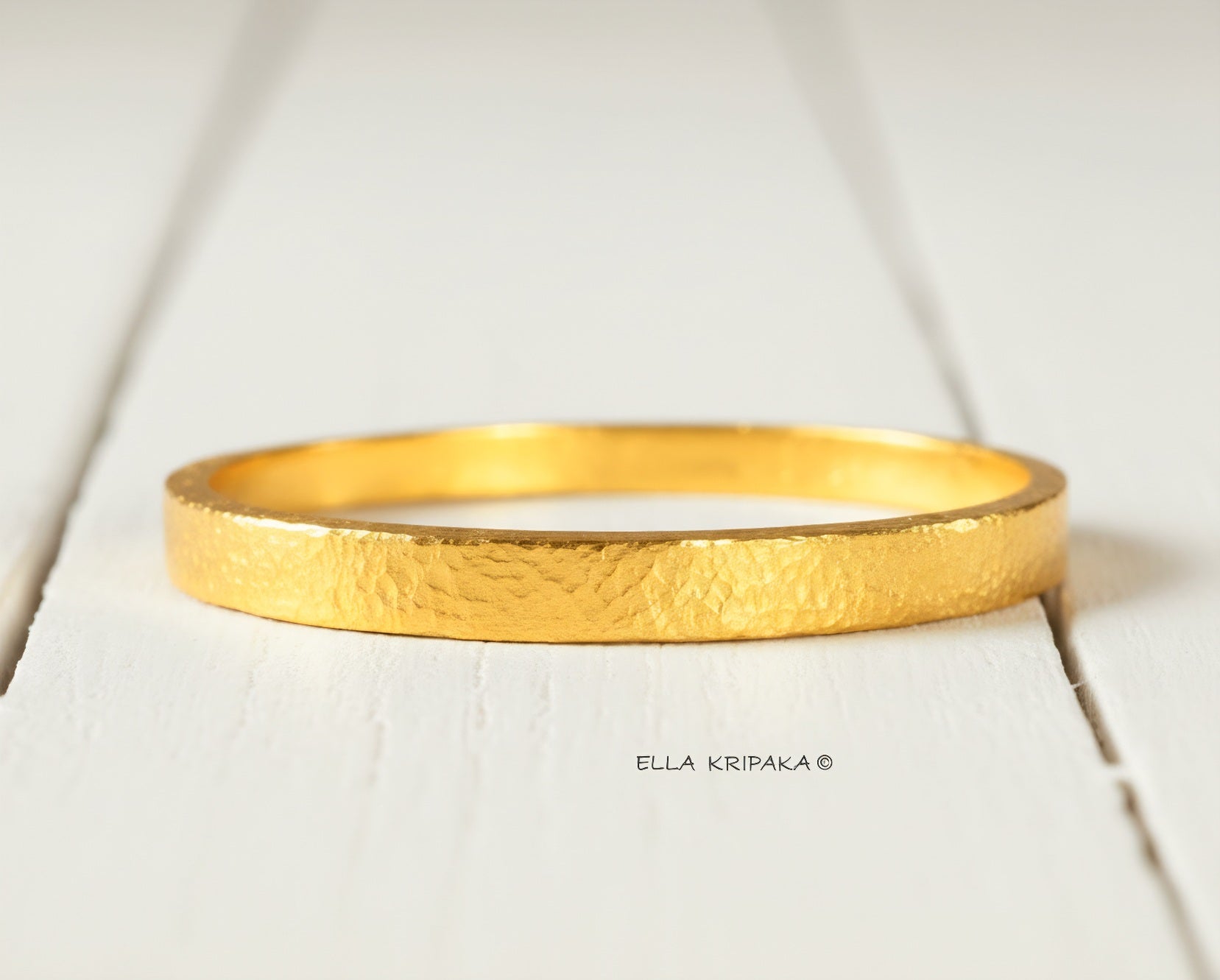 Hammered 24k 9999 gold bangle thick bracelet Ancient Rome style on a white wooden background, artist-jeweler name 'Ella Kripaka' branding