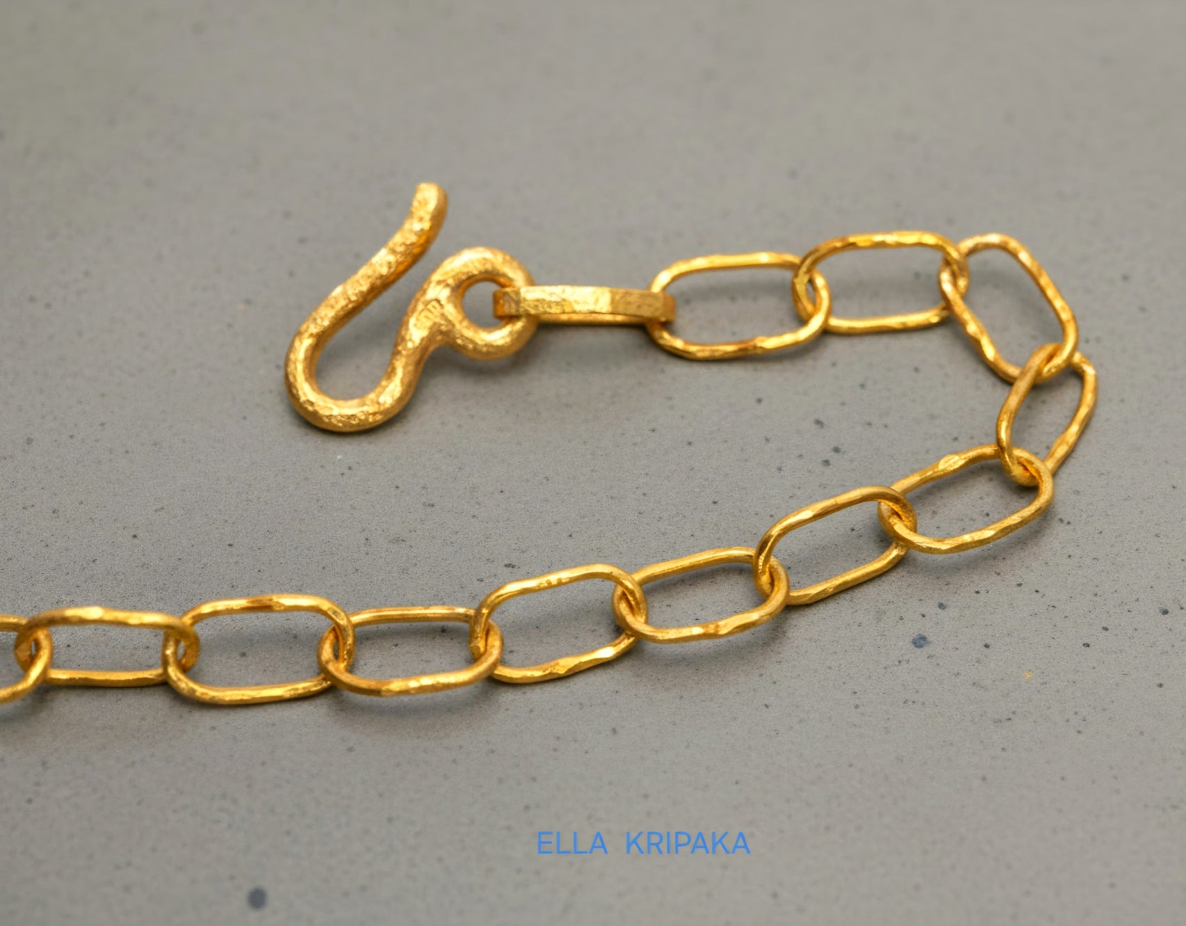 22k gold hammered chain with thick hook clasp on grey concrete background, artist-jeweler's name 'Ella Kripaka' branding