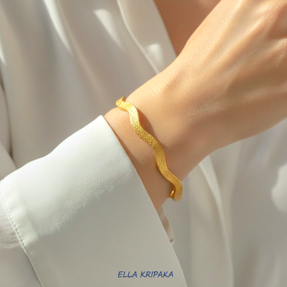 24k gold hammered bracelet bangle with wavy design is wearing on model's wrist and on blurred white shirt background, branded 'Ella Kripaka'