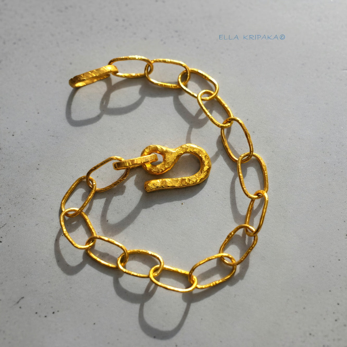 22k gold hammered chain bracelet with thick hook clasp on grey concrete background, artist-jeweler's name 'Ella Kripaka' branding