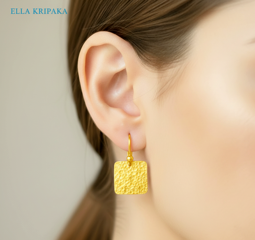 Hammered 24k gold, pure 9999 gold square earring with shiny forged gold textures worn on a woman and with light grey background, artist-jeweler 'Ella Kripaka' branding