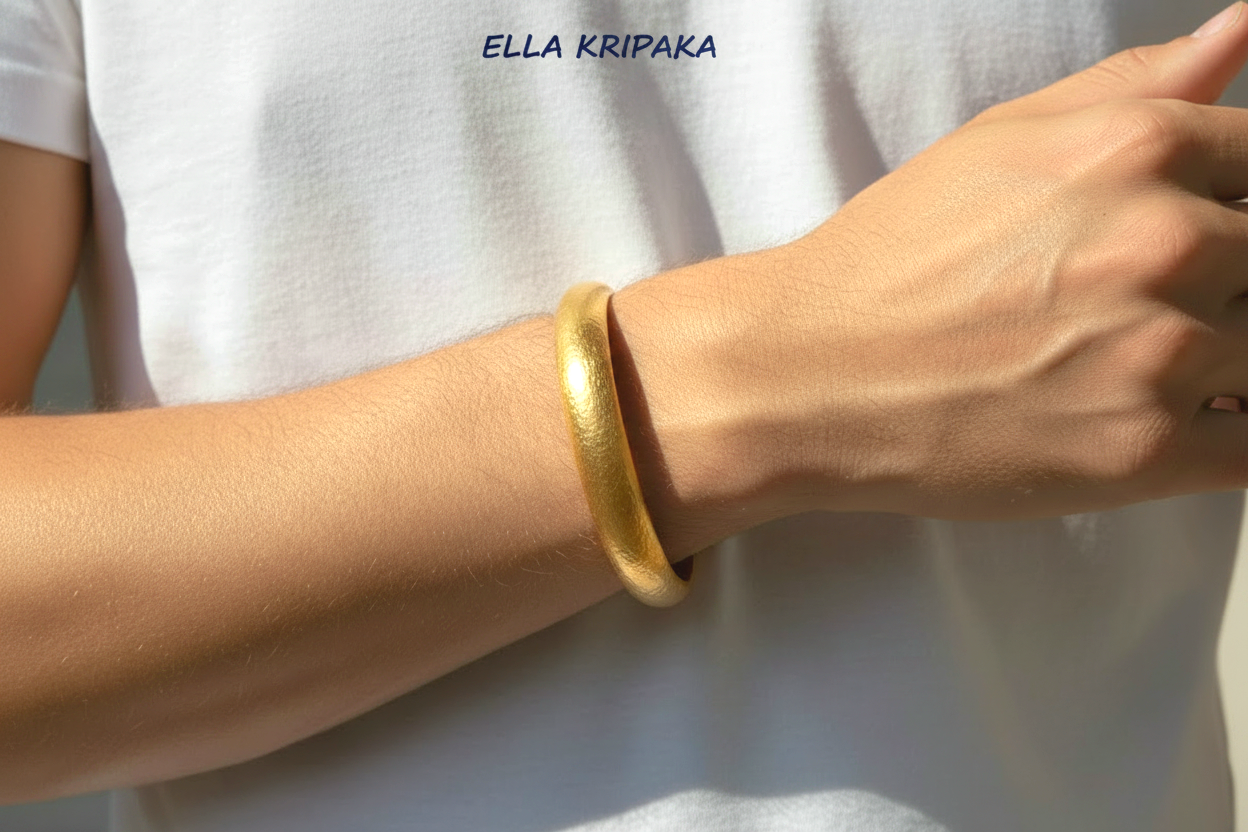 Hammered 24k gold bangle thick bracelet with rounded edges is worn by a man on wrist and on white t-shirt background, artist-jeweler 'Ella Kripaka' branding