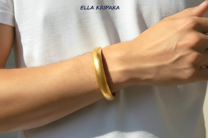 Hammered 24k gold bangle thick bracelet with rounded edges is worn by a man on wrist and on white t-shirt background, artist-jeweler 'Ella Kripaka' branding