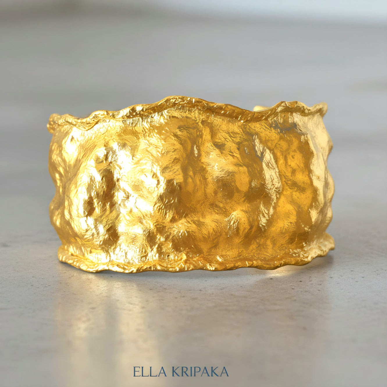 Organic 24k gold hammered bracelet cuff with a textured surface on a grey concrete background, branded 'Ella Kripaka'