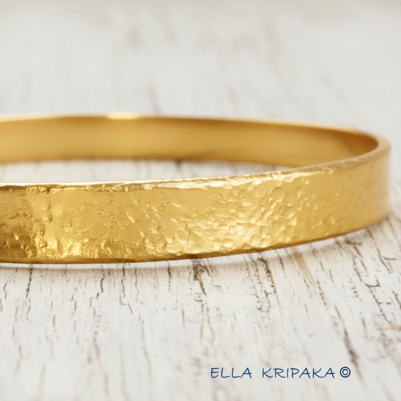 24k 9999 gold hammered Ancient Rome style bracelet bangle on white rustic wooden surface, indicating it is a custom-made jewelry with creator's name 'Ella Kripaka'