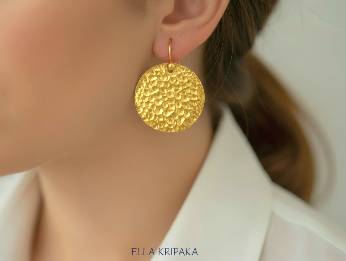 Organic hammered 24k gold, pure 9999 gold disk earring with shiny forged gold textures worn on a woman and with light grey background, artist-jeweler 'Ella Kripaka' branding