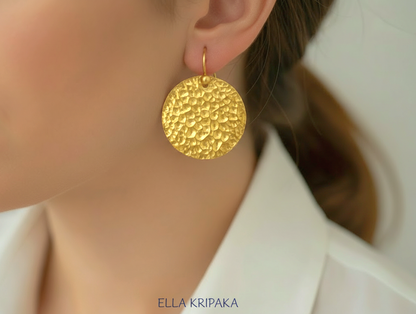 Organic hammered 24k gold, pure 9999 gold disk earring with shiny forged gold textures worn on a woman and with light grey background, artist-jeweler 'Ella Kripaka' branding
