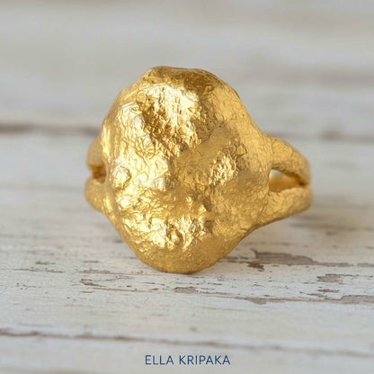 Organic 24k gold convex signet ring with textured uneven design on a light rustic wooden surface, branded 'Ella Kripaka'