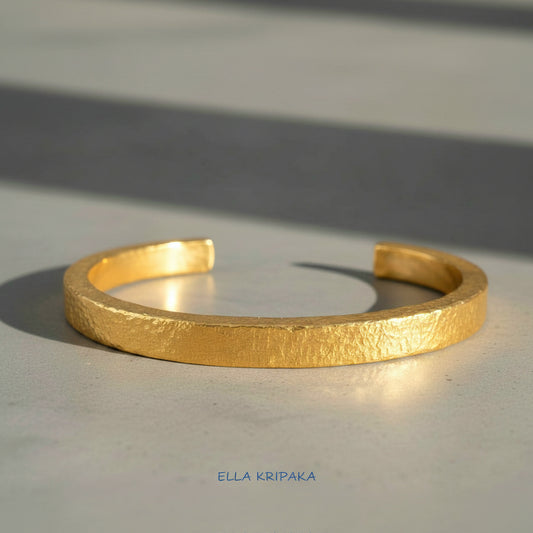 Thick hammered 24k gold shiny bracelet cuff on a light  grey concrete surface, with artist 'ELLA KRIPAKA' branding