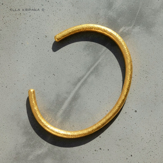 Hammered 24k 9999 gold cuff textured bracelet on a grey concrete background, artist-jeweler name 'Ella Kripaka' branding
