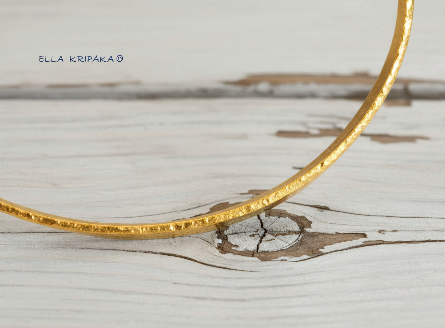 24k 9999 gold hammered Ancient Rome style bracelet bangle on white rustic wooden surface, indicating it is a custom-made jewelry with creator's name 'Ella Kripaka'