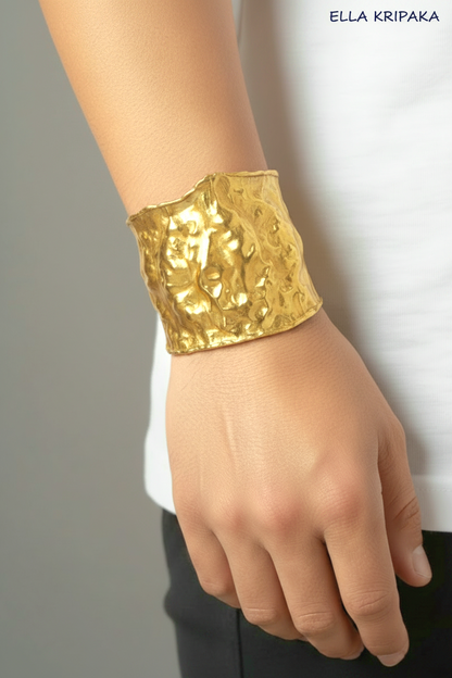 Hammered 24k gold 65mm cuff bracelet with convex uneven textures is worn by a man on wrist and on grey background, and with 'Ella Kripaka' branding
