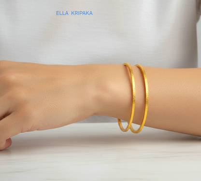 22k gold hammered two thick bangles bracelets Ancient Rome style worn by a woman on wrist and on a white wooden surface, artist-jeweler 'Ella Kripaka' branding