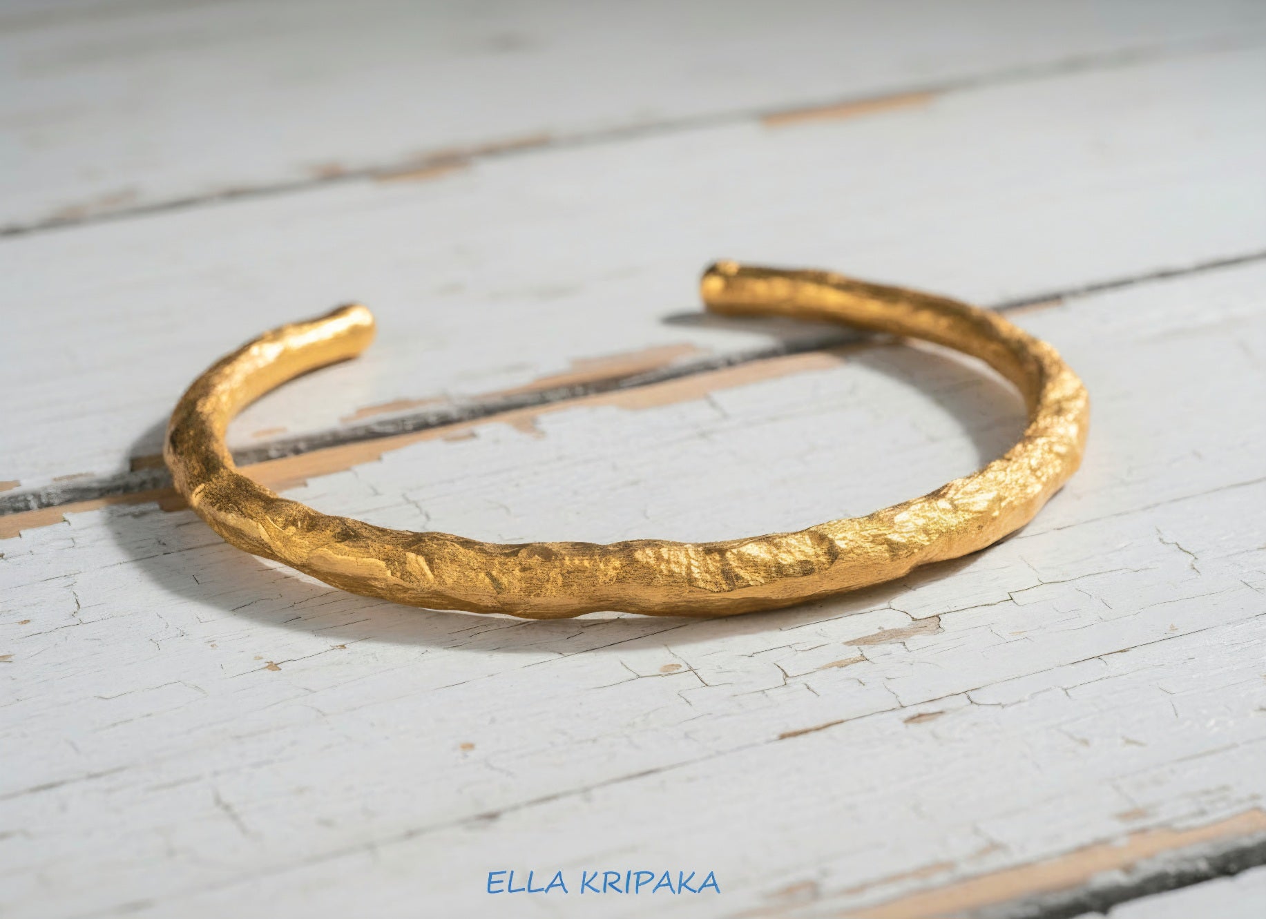 Organic hammered 24k 9999 gold bracelet uneven cuff on a old white old wooden background, branding artist-jeweler name 'Ella Kripaka'