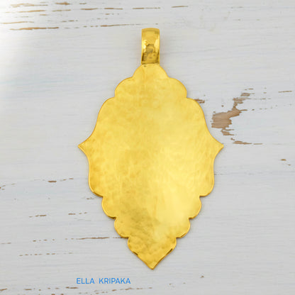 Hammered 24k gold Byzantine ornament pendant on a white rustic wooden surface with 'Ella Kripaka' branding