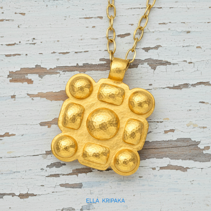 Hammered 24k gold square pendant with convex pattern and with chain on a white rustic wooden surface with 'Ella Kripaka' branding
